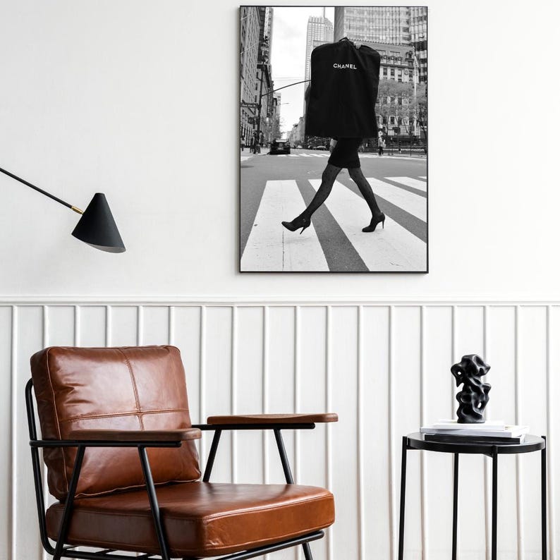 May include: Black and white framed photograph of a person crossing a crosswalk in a city. The person is wearing a black dress and heels, carrying a garment bag with the word "CHANEL" printed on it. The background features buildings and a street.
