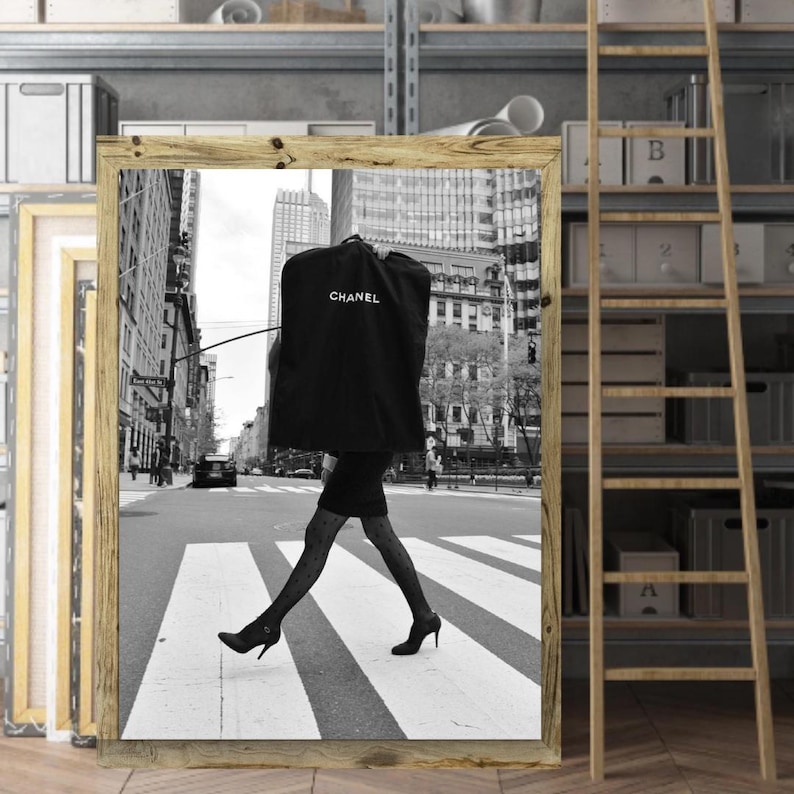May include: Black and white framed print of a person crossing a city street in high heels, carrying a garment bag. The garment bag is black with the word "CHANEL" printed in white. The background shows buildings and a crosswalk.