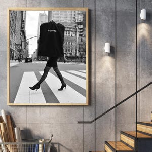 May include: Black and white framed print of a person crossing a crosswalk in a city. The person is wearing a black dress, patterned stockings, and heels, carrying a garment bag with the Chanel logo.