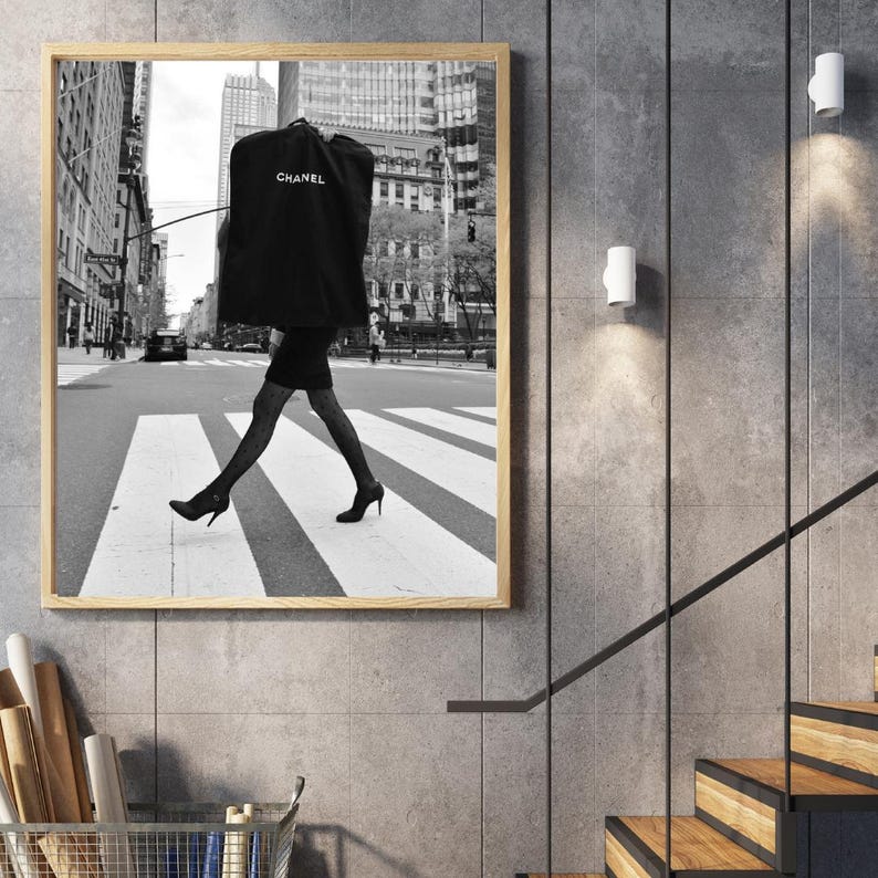 May include: Black and white framed print of a person crossing a crosswalk in a city. The person is wearing a black dress, patterned stockings, and heels, carrying a garment bag with the Chanel logo.