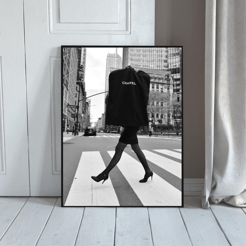 May include: Black and white framed print of a person crossing a crosswalk in a city. The person is wearing a black dress, patterned stockings, and heels, carrying a garment bag with the Chanel logo.