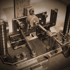 Könnte beinhalten: Nahaufnahme einer Vintage-Metallmaschine mit verschiedenen mechanischen Komponenten, darunter Schrauben, Federn und Hebel. Die Maschine hat einen Sepia-Ton, der eine antike oder industrielle Ästhetik suggeriert. Das Design ist komplex und detailliert.