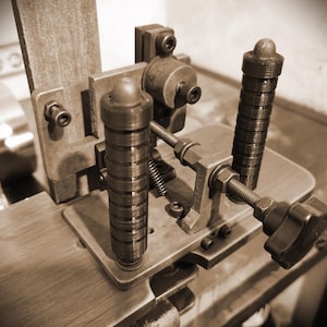 May include: A sepia-toned image of a vintage metal machine. It features cylindrical handles, screws, and a wooden base. The intricate mechanical device has various components, showcasing its detailed construction.