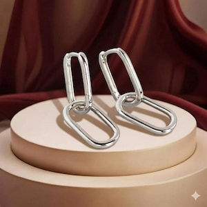 Puede incluir: Un par de pendientes plateados con forma de eslabones de cadena. Cada pendiente presenta dos eslabones ovalados conectados. Los pendientes se exhiben sobre una superficie rosa claro con un fondo de tela burdeos.