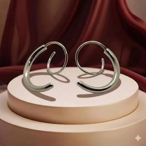 May include: A pair of silver hoop earrings. The earrings are a curved, semi-circular design with a smooth, polished finish. They are displayed on a light pink, circular platform against a backdrop of burgundy fabric.