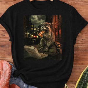 May include: Black t-shirt featuring a raccoon wearing a cap and eating pizza in a city setting. The raccoon is sitting on a wooden crate next to an open pizza box. The background shows buildings and streetlights.
