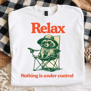 May include: White t-shirt with a graphic of a raccoon wearing sunglasses and a hat, relaxing in a folding chair. The word "Relax" is printed above the raccoon in orange, with "Nothing is under control" below.