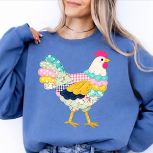 May include: A blue sweatshirt featuring a colorful patchwork chicken design. The chicken has a white body, a pink comb, and a yellow beak. Its tail and wings are made of various patterned fabrics, including polka dots, floral prints, and gingham.