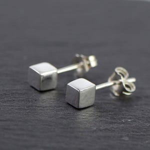 May include: A pair of silver cube stud earrings. Each earring features a small, square-shaped design with a post and butterfly backing. The earrings are displayed on a dark, textured surface.