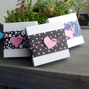 May include: Three white gift boxes with decorative bands and pink heart accents. One box has a black and white floral band, another has a black polka dot band, and the third has a blue floral band. Each box is tied with pink string.