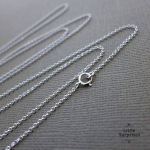 May include: Close-up of several delicate silver chain necklaces with a lobster clasp closure. The chains are displayed on a gray surface. The image is well-lit, highlighting the reflective quality of the metal. The necklaces are a simple, elegant design.