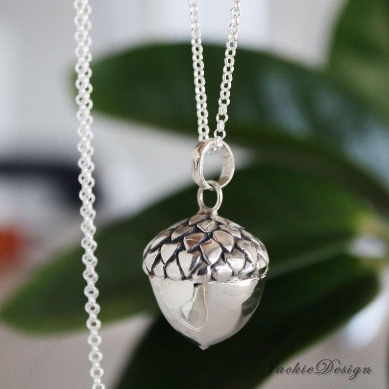 May include: A silver acorn pendant necklace. The pendant features a detailed acorn design with a textured cap and a smooth, rounded body. The necklace chain is delicate and silver, hanging against a blurred green background.