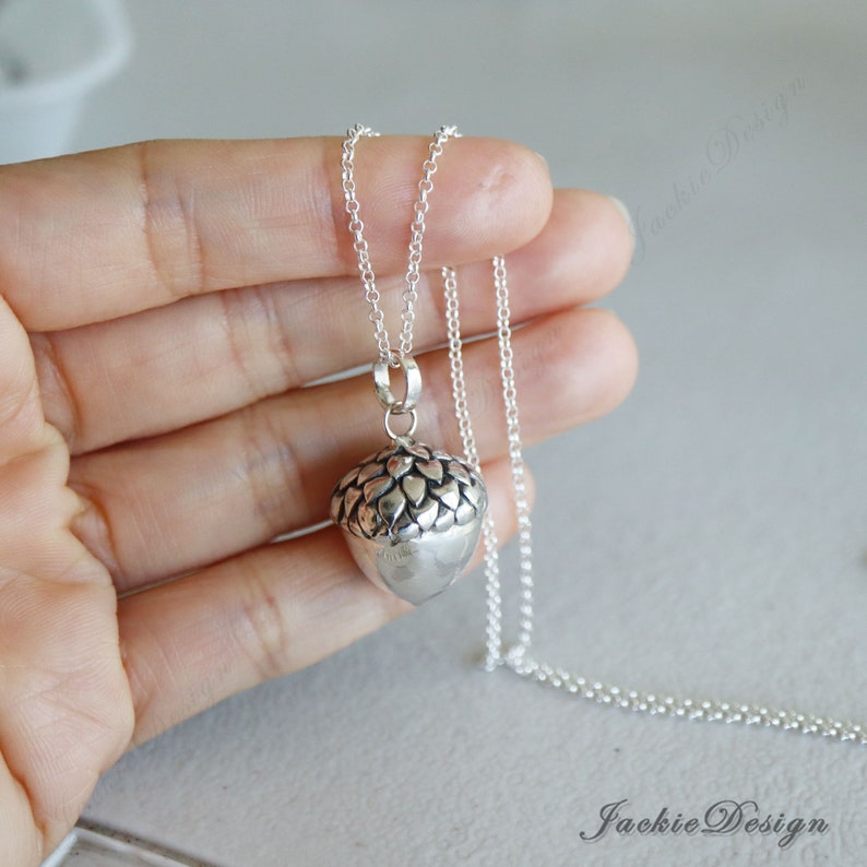 May include: A silver acorn pendant necklace. The pendant is a detailed acorn design with a textured cap and smooth body. The necklace chain is delicate and silver, creating a simple, elegant look. The jewelry is displayed against a neutral background.