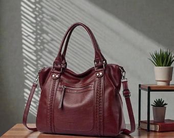 May include: A burgundy leather handbag with a top handle and a detachable shoulder strap. The bag has a front zipped pocket and braided detailing on the sides. It sits on a wooden table.