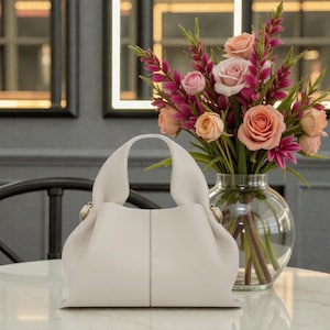 May include: A cream-colored handbag with a rounded handle sits on a marble table. The bag has a soft, gathered design and gold-colored hardware. A vase of pink and peach roses and greenery is in the background.