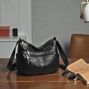 May include: Black shoulder bag with multiple zipped compartments and a long adjustable strap. The bag has a textured appearance and a classic design. The bag is on a wooden surface.