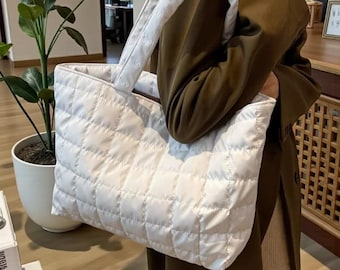 Könnte beinhalten: Eine weiße, gesteppte Tragetasche mit einem Rautenmuster. Die Tasche hat zwei weiße Griffe. Die Tasche wird von einer Person gehalten, die einen langen olivgrünen Mantel trägt.