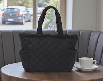 May include: A black quilted tote bag with two exterior pockets and two handles. The bag is on a wooden table next to a white teacup and saucer. The bag features a diamond pattern.
