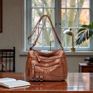 Puede incluir: Un bolso de hombro de cuero marrón con múltiples bolsillos con cremallera y un detalle lateral trenzado. El bolso tiene una correa larga ajustable y está sobre una mesa de madera. El fondo incluye una ventana, una lámpara y una planta.