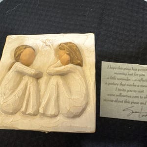 Könnte beinhalten: Eine cremefarbene Reliefskulptur mit zwei eng beieinander sitzenden Figuren, deren Köpfe sich berühren. Zwischen ihnen befindet sich ein kleiner Stern. Eine kleine Karte mit Text liegt daneben.