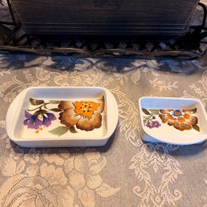 May include: Two white ceramic dishes with floral designs. The larger dish is rectangular, and the smaller one is square. The floral pattern includes purple, orange, and brown flowers with green leaves. A decorative metal tray is in the background.