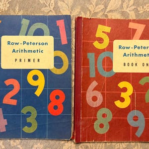 May include: Two vintage arithmetic textbooks titled Row-Peterson Arithmetic Primer and Row-Peterson Arithmetic Book One. The blue primer features colorful numbers on a grid, while the red book displays similar numbers on a matching grid pattern.