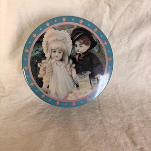 May include: Round tin with a blue border featuring a decorative illustration of two antique dolls. One doll wears a large hat and white dress, the other a black coat and hat. A pitcher and small cakes are also visible.