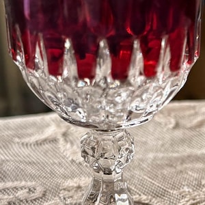 May include: A crystal wine glass filled with a deep red liquid. The glass has a clear, faceted bowl and a decorative stem. The liquid appears to be wine, and the glass is set on a textured surface.