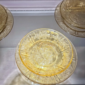 May include: A collection of vintage amber-colored glass dishes. The set includes plates and bowls with a floral embossed design. The glass has a warm, golden hue, and the dishes are arranged on a reflective surface, showcasing their intricate details.