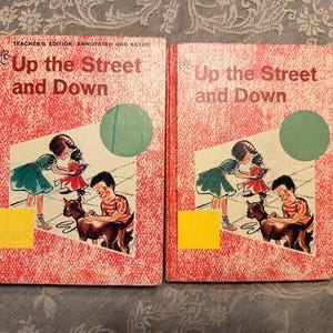 May include: Two identical hardcover books titled Up the Street and Down with red and white speckled covers. The front covers feature an illustration of a child in a green dress and a child in a striped shirt with a dog, plus a yellow square and green circle.