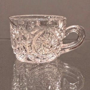 May include: A clear glass cup featuring a pressed pattern of circular motifs and geometric starburst designs. The cup has a thick, curved handle and a textured surface that catches the light, creating a multifaceted appearance across the exterior.