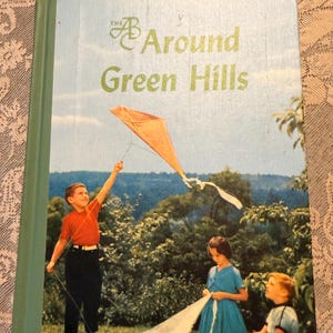 May include: A hardcover book titled The Around Green Hills with a green spine. The cover features an illustration of a child in a red shirt flying an orange kite, while another child in a blue dress and a child in a white shirt sit nearby.