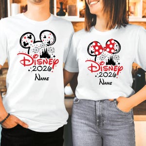 K&ouml;nnte beinhalten: Wei&szlig;e T-Shirts mit einem Disney-Motiv. Ein Shirt zeigt einen schwarzen Mickey Mouse-Kopf mit roten Sternen, Feuerwerk und dem Text "Disney 2026 Name" in Rot. Das andere Shirt hat eine rote Schleife mit Punkten.