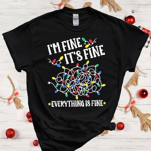 May include: A black short-sleeve t-shirt featuring the white text I'M FINE IT'S FINE and EVERYTHING IS FINE. The design includes a graphic of a tangled mess of white Christmas light wires with colourful red, blue, green, and yellow light bulbs.