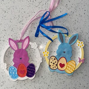 May include: Two Easter bunny decorations. One is purple with a white frame, decorated with eggs in blue, orange, and purple. The other is blue with a yellow frame, decorated with eggs in brown, orange, and yellow. Both have ribbon hangers.
