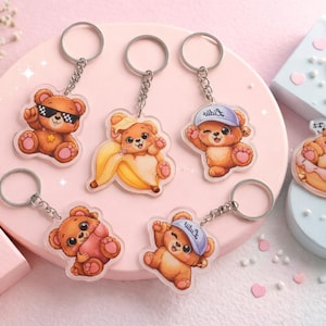 Könnte beinhalten: Eine Sammlung von Acryl-Schlüsselanhängern mit Cartoon-Teddybären in verschiedenen Posen. Die braunen Bären haben Accessoires wie Sonnenbrillen, eine Banane, eine Baseballkappe und einen Donut. Jeder Schlüsselanhänger hat einen silbernen Ring.