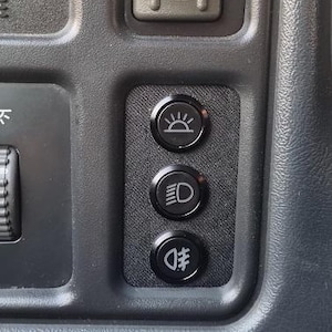 May include: Close-up of a vehicle dashboard with buttons. The buttons include 4 HI, 4 LO, a sun symbol, a headlight symbol, and a fog light symbol. The buttons are black and gray, set against a dark gray background.