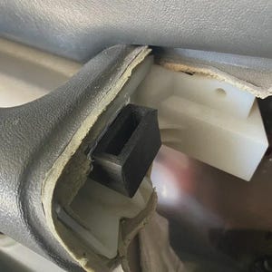 May include: Close-up of a car door panel with a black rectangular foam insert. The panel is gray with a textured surface, and the foam insert is positioned within a recessed area. A white plastic component is visible.