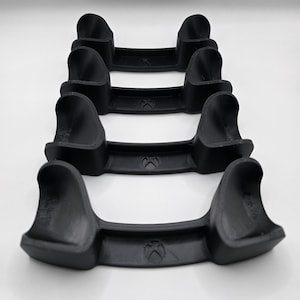 May include: Four black game controller stands arranged in a vertical stack. Each stand has a curved design with a central Xbox logo. The stands are made of a matte black material and are designed to hold game controllers.