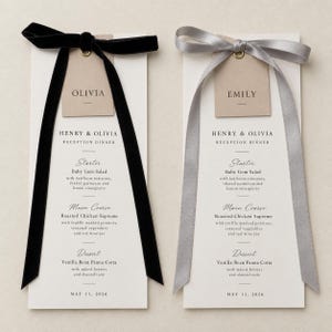 Peut inclure: Deux menus de dîner de réception blancs et hauts avec du texte noir. L'un présente un ruban de velours noir et une étiquette OLIVIA, l'autre un ruban de satin argenté et une étiquette EMILY. Ils listent des plats comme la salade Baby Gem, le poulet rôti et la panna cotta à la vanille pour le 11 mai 2026.