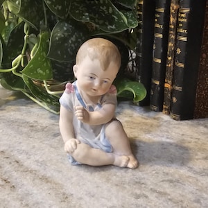 Może przedstawiać: Porcelanowa figurka siedzącego niemowlęcia ubranego w biały strój z jasnoniebieskim wykończeniem i małymi różowymi akcentami na ramionach. Niemowlę ma jasnobrązowe włosy, niebieskie oczy i siedzi z jedną ręką uniesioną do góry.