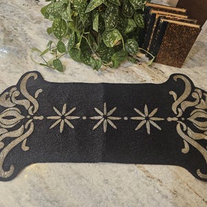 May include: A rectangular black table runner featuring intricate gold beaded scrollwork on both ends and a central row of three gold beaded starburst motifs. The runner has a scalloped edge design and is crafted with a dense, uniform beaded texture.