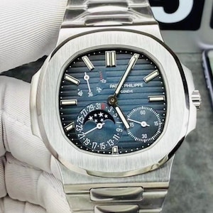 May include: A silver-toned stainless steel wristwatch with a blue horizontal-striped dial. The watch features a moon phase indicator, a date sub-dial, and a power reserve display. The text Patek Philippe Geneve is printed on the watch face.