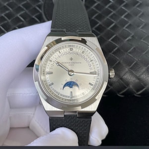 May include: A silver-toned wristwatch with a black rubber strap. The watch face features a moon phase indicator and the brand name "VACHERON CONSTANTIN GENEVE". The watch has a round face with a date display and a textured black strap.