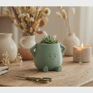 May include: A sage green ceramic planter shaped like a person with arms akimbo and a sad face. The planter holds a small succulent plant. The scene includes a lit candle, vases with dried flowers, and a pair of scissors on a wooden table.