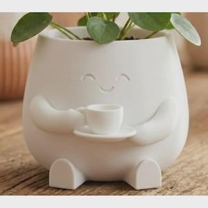 May include: A white ceramic planter with a smiling face, arms, and legs, holding a small teacup and saucer. The planter contains a green plant with round leaves. The design is whimsical and decorative.