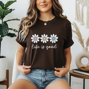 May include: A brown t-shirt featuring three daisy flowers with pink, yellow, and blue centers. The words "life is good" are written in a cursive font below the flowers. The shirt is a casual, everyday wear item.