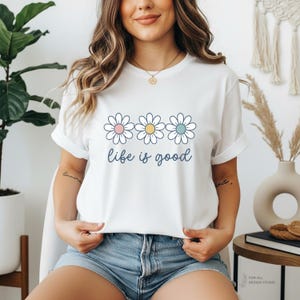 May include: White t-shirt with a daisy graphic and the phrase "life is good" in a script font. The daisy graphic features three flowers with pink, yellow, and blue centers. The shirt has rolled-up sleeves and is worn with blue denim shorts.