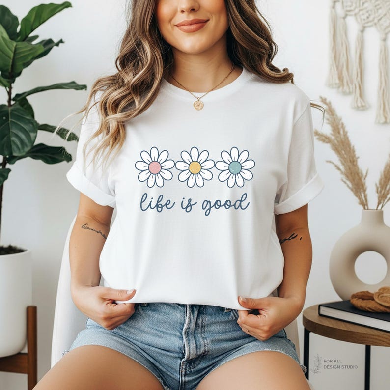 May include: White t-shirt with a daisy graphic and the phrase "life is good" in a script font. The daisy graphic features three flowers with pink, yellow, and blue centers. The shirt has rolled-up sleeves and is worn with blue denim shorts.