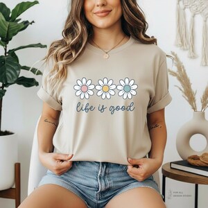 May include: Beige t-shirt with a daisy graphic and the phrase "life is good" in blue script. The daisy design features three flowers with pink, yellow, and blue centers. The shirt is made of a soft material.